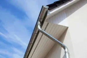 A metal gutter and downspout attached to the corner of a house with white siding under a bright blue sky.