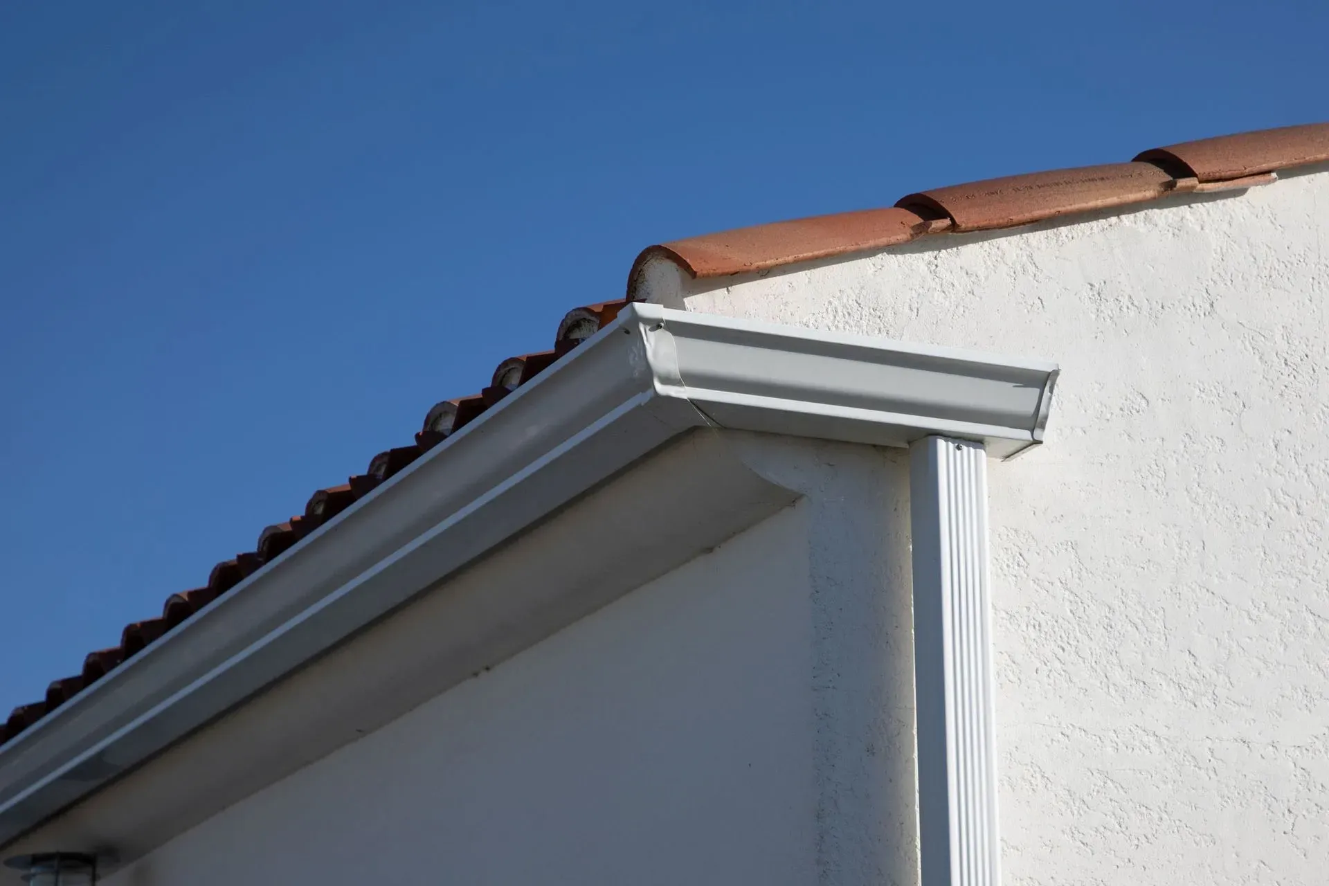 White rain gutters and a downspout installed on the white exterior wall of a building with a red-tiled roof.