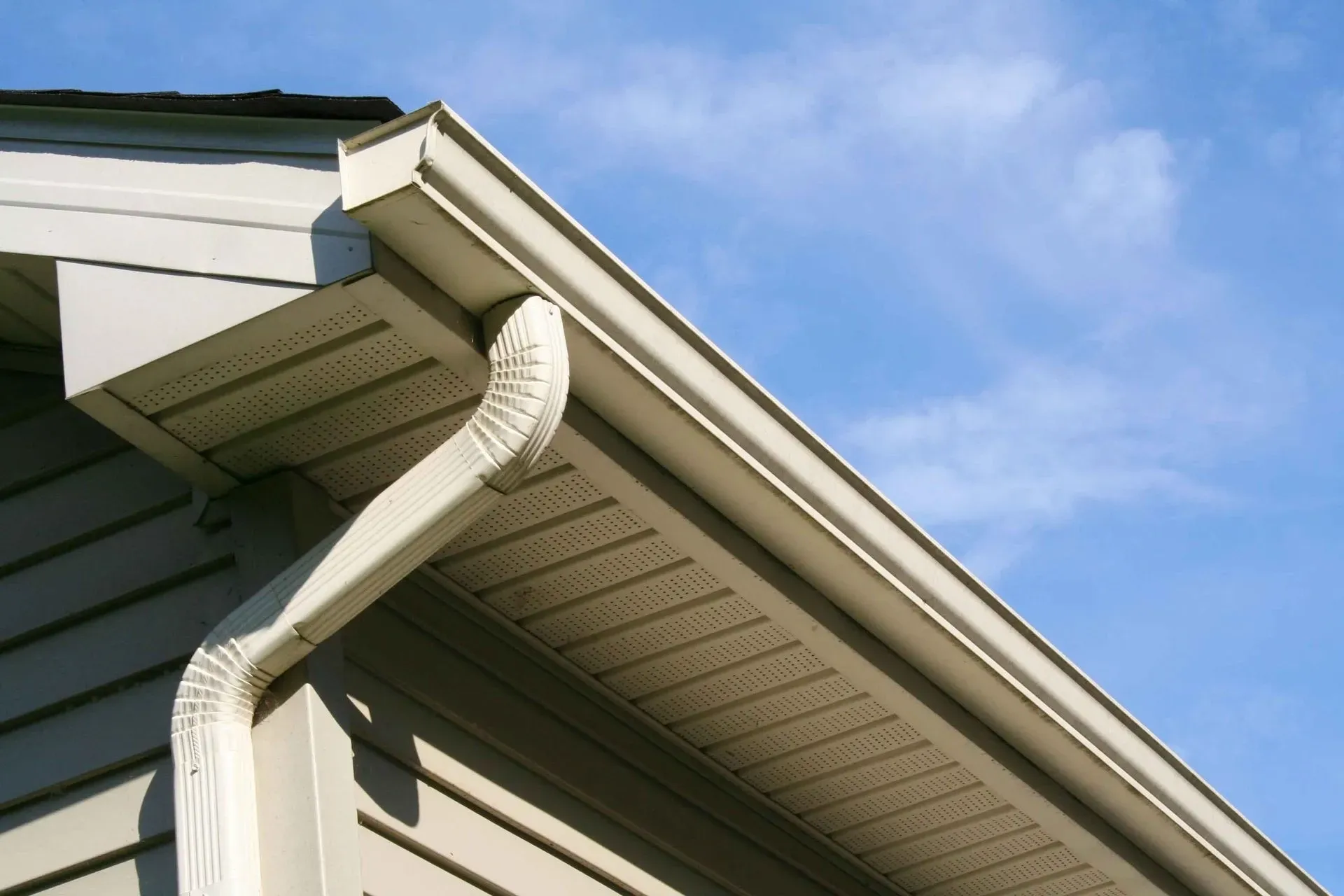Beige vinyl siding and soffit with a white gutter and downspout under a blue sky.