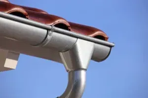 A metallic gutter installed beneath brown curved roof tiles against a clear blue sky.