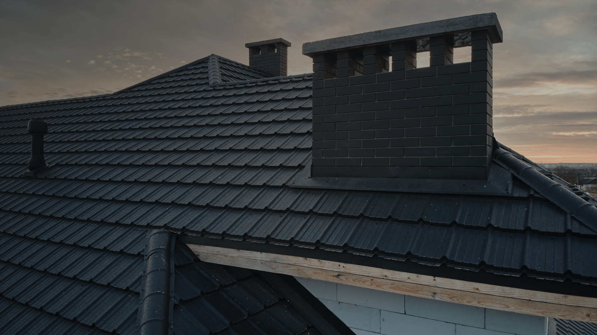 A dark tiled roof featuring a brick chimney and a small vent, set against a cloudy sky at sunset.