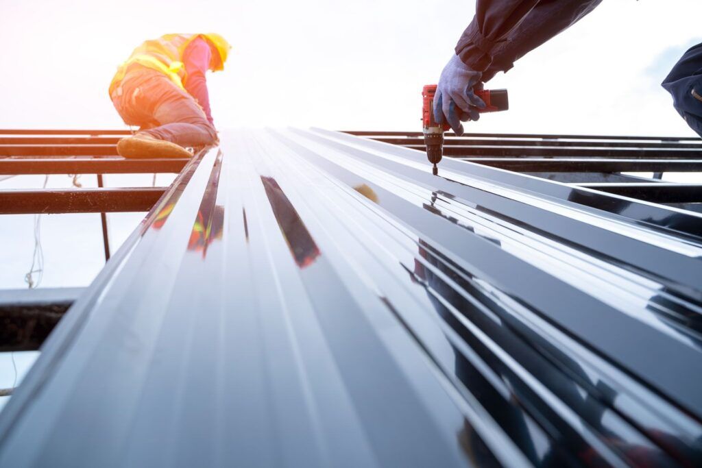 Construction workers installing shiny metal roofing panels with a drill.
