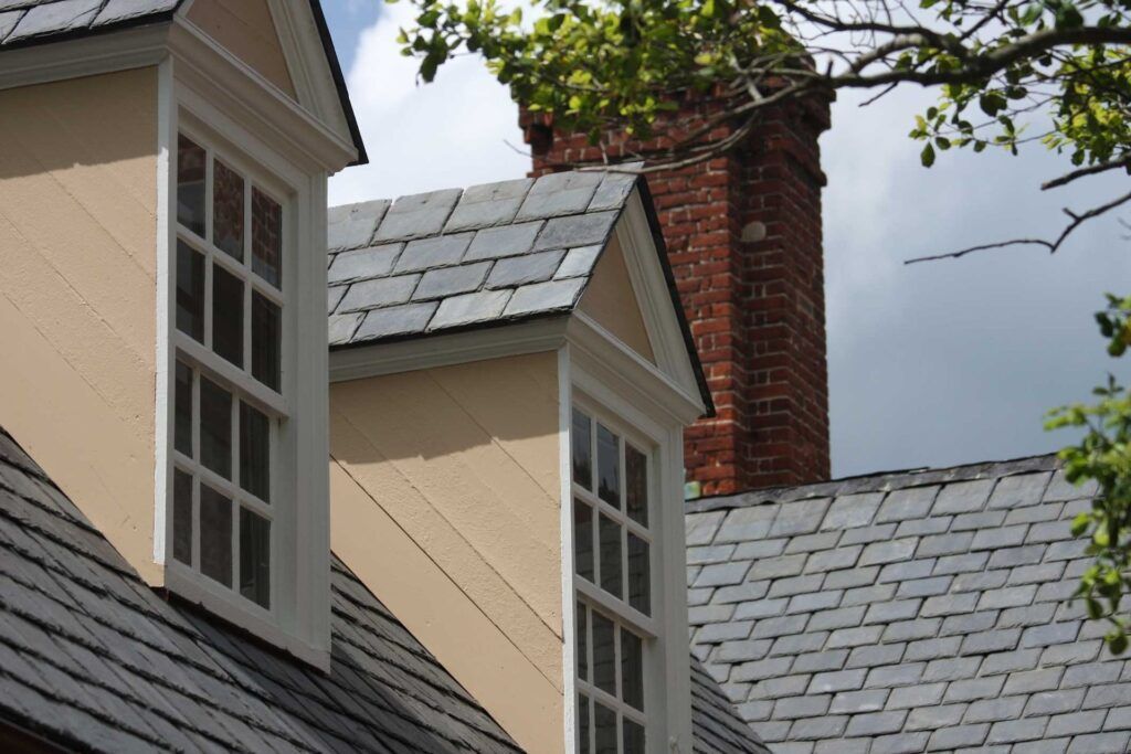 Two beige dormer windows with white trim and slate roofs, set against a red brick chimney and a cloudy sky.