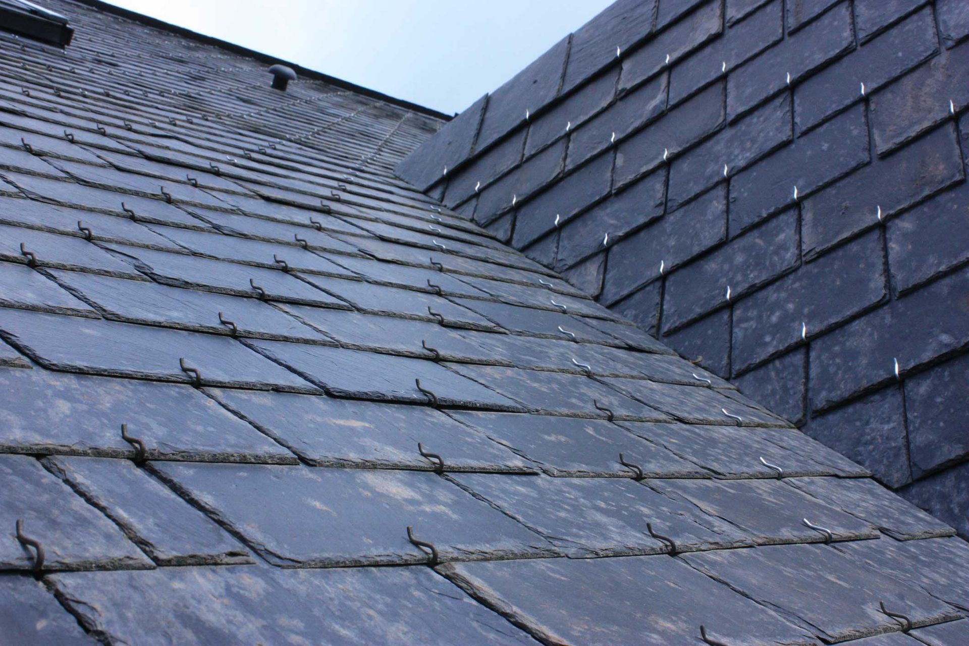 A close-up view of a dark grey slate roof showing overlapping tiles and metal snow guard hooks.