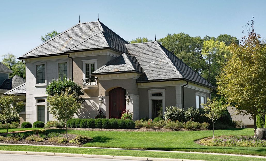 Two-story gray house with dark roof, front porch, and manicured lawn on a sunny suburban street