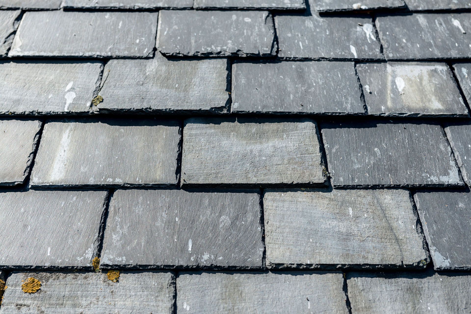 A close-up view of overlapping gray, rectangular roof slates with weathered textures and small patches of lichen.