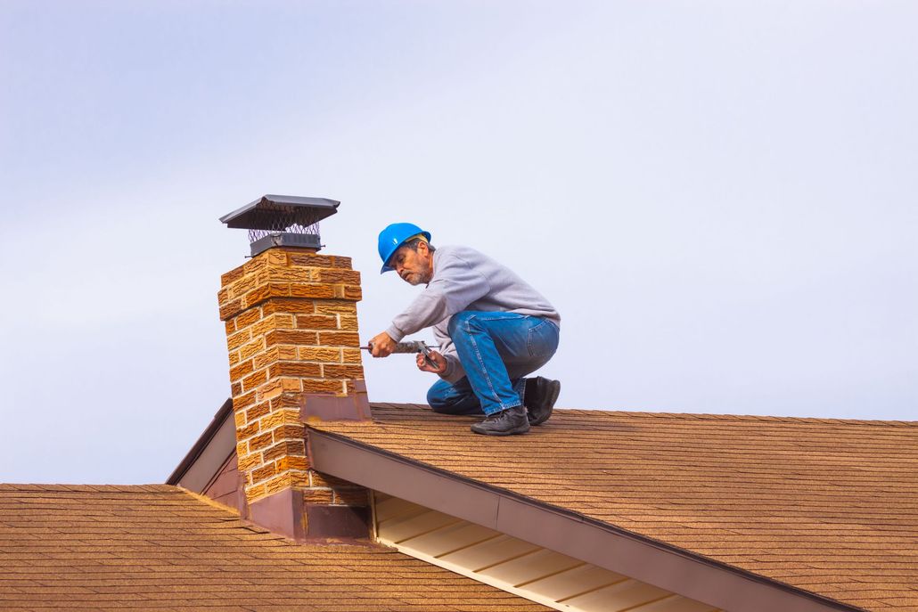 A worker in a blue hard hat kneels on a shingled roof, applying sealant to the base of a brick chimney.