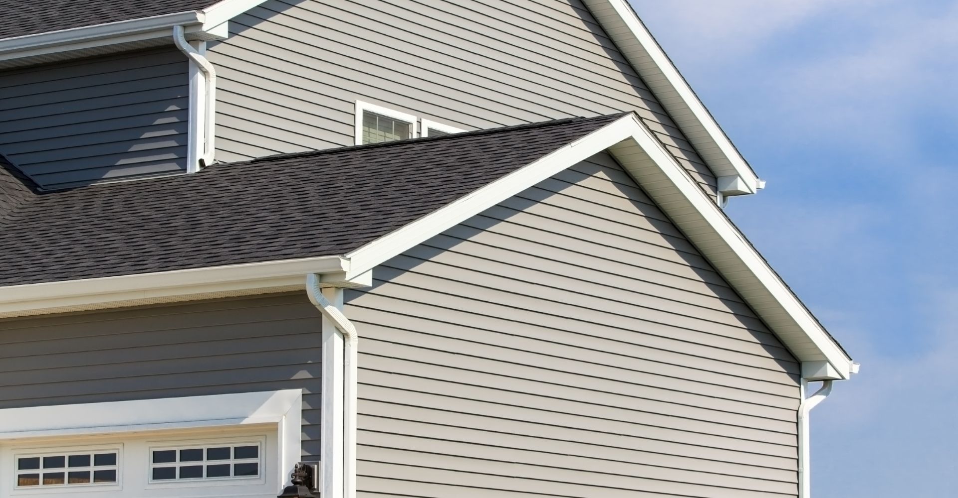 The upper portion of a house exterior featuring grey horizontal siding, a dark shingled roof, and white trim.