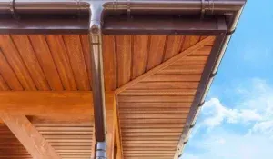 A close-up view of the underside of a wooden roof overhang and a dark brown downspout against a bright blue sky.