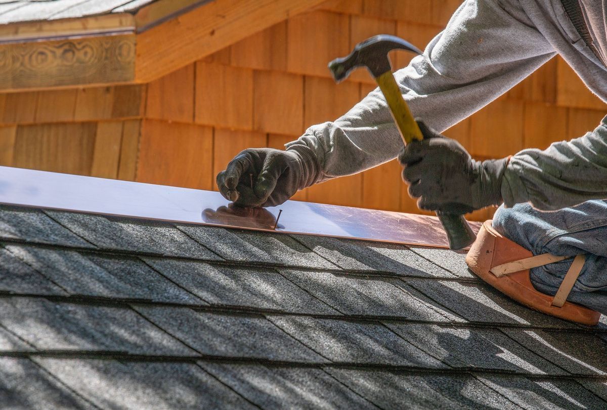 A person wearing gloves hammers a piece of copper flashing onto a dark shingled roof.