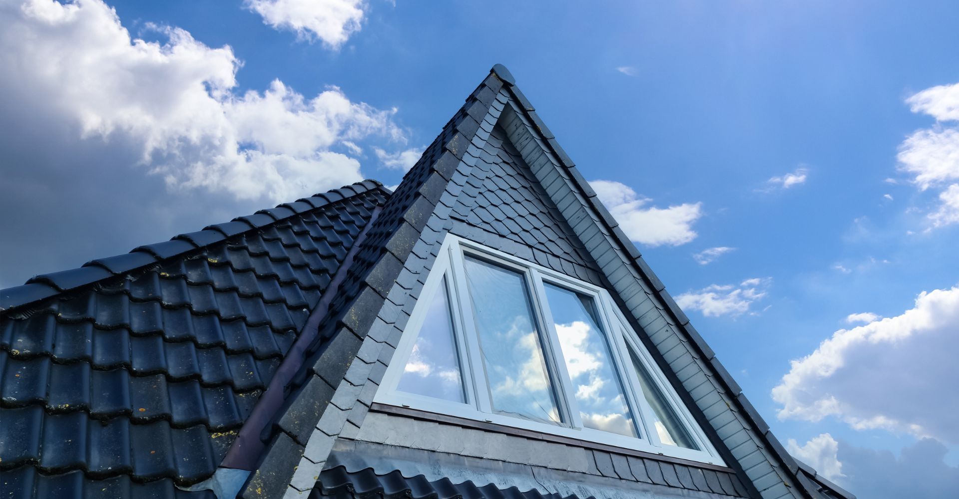 A triangular dormer window set into a dark tiled roof against a bright blue sky with scattered white clouds.