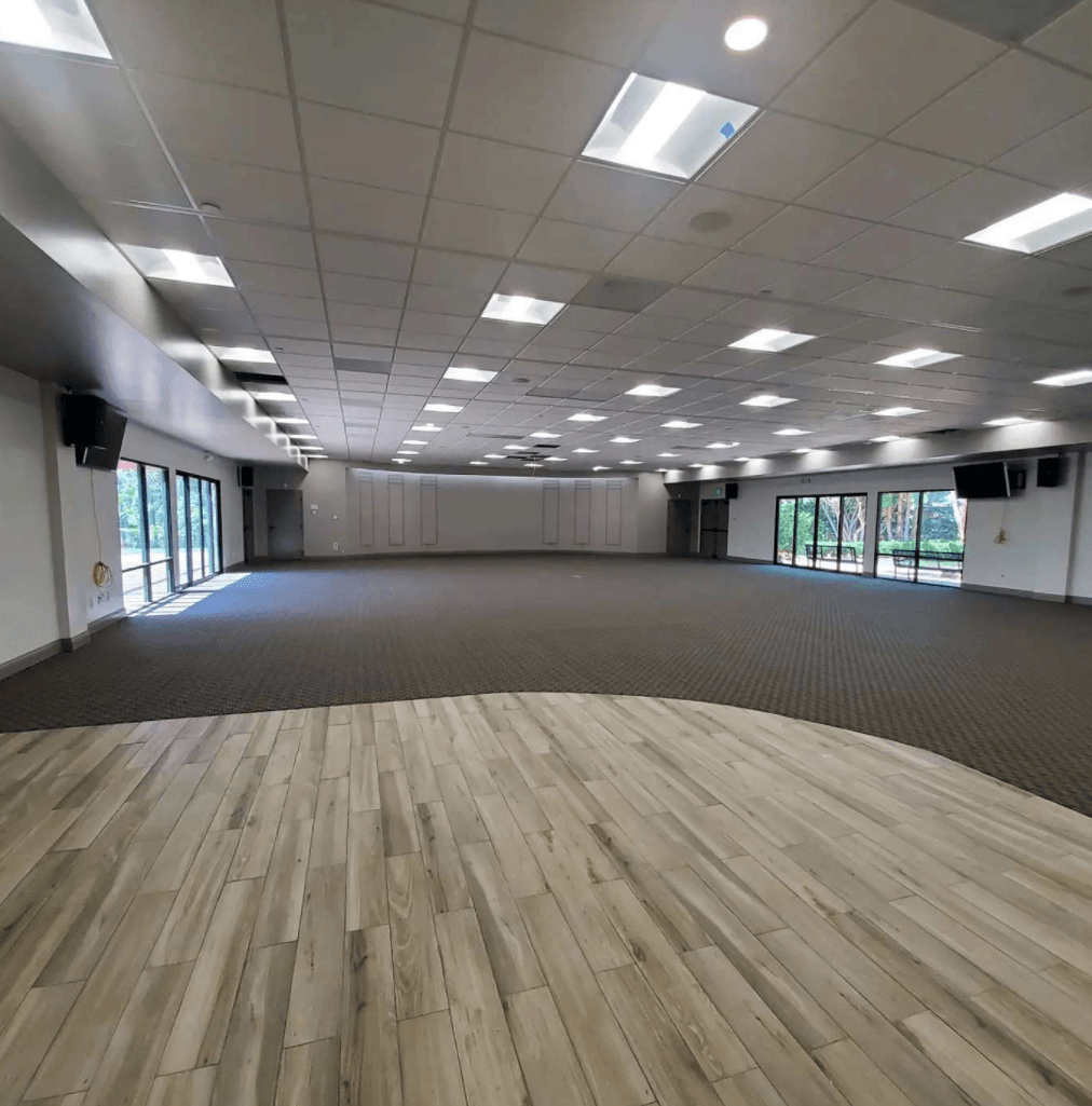 A large, empty event hall featuring a light wood-plank floor in the foreground, dark carpeting, and recessed ceiling lights.