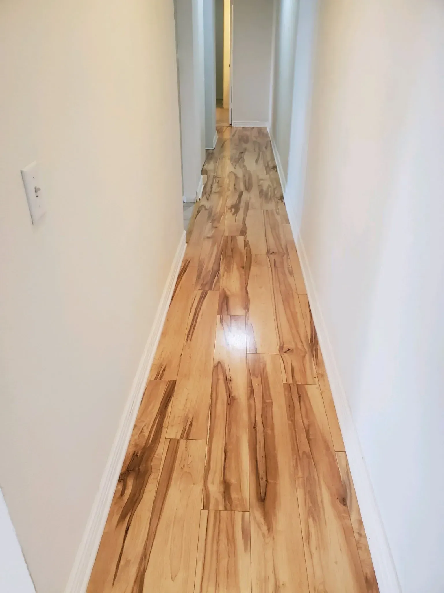 A long, narrow hallway with light-colored, wood-grained flooring and white walls leading to a bright doorway.