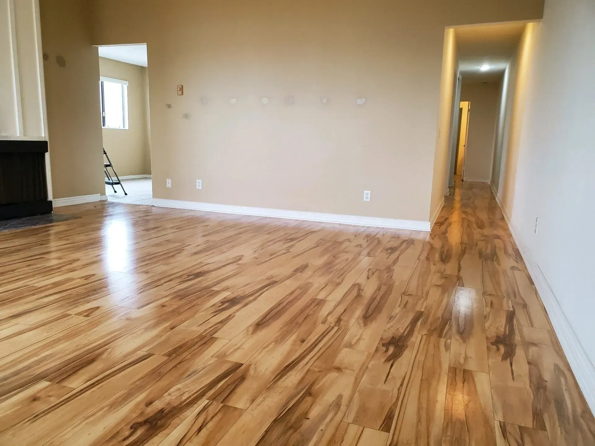 A brightly lit, empty living room with glossy, light-colored wood flooring and tan walls, leading to a long hallway.