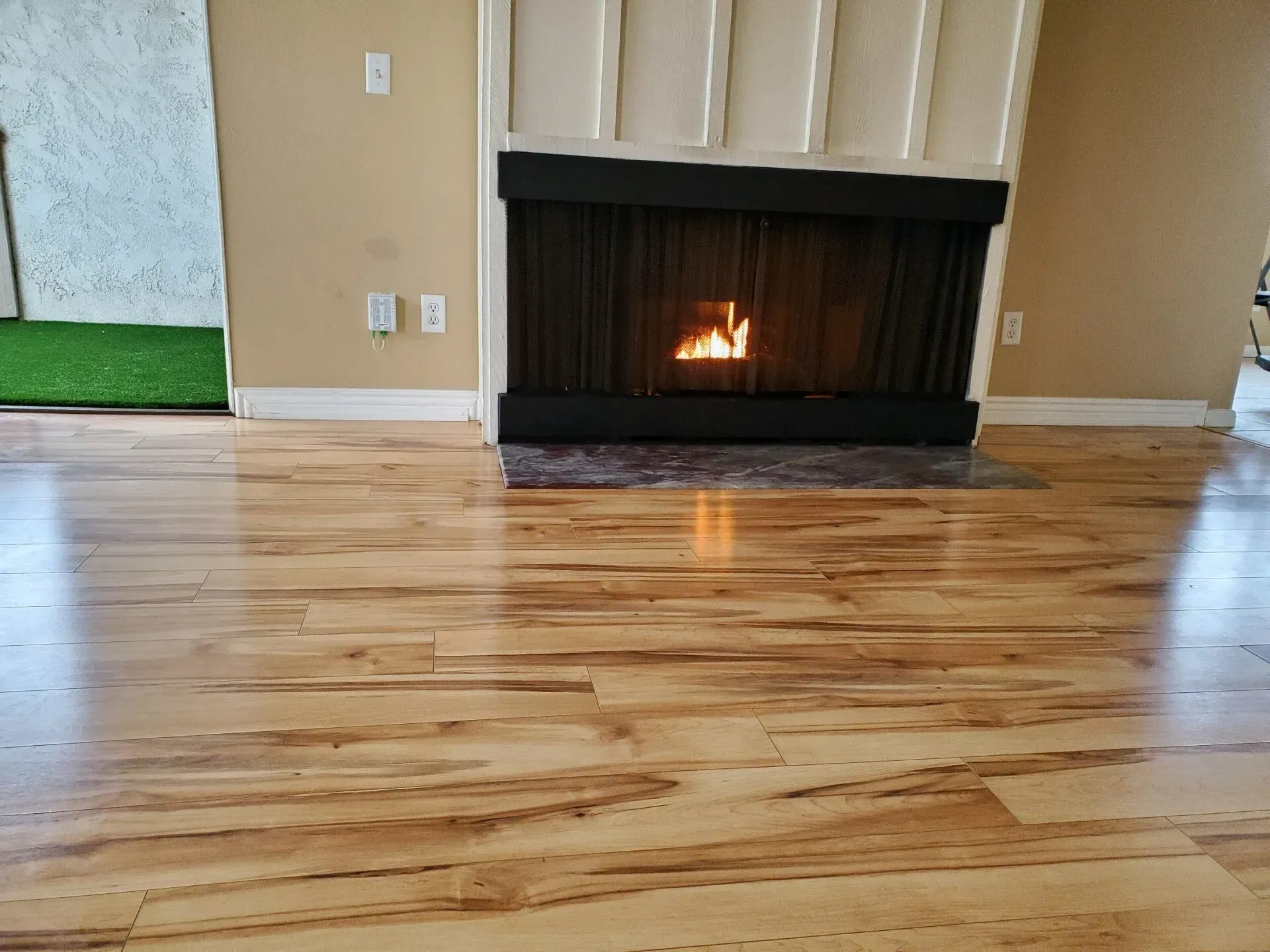 A fireplace with a lit fire set against a paneled wall above light-colored, multi-toned wood flooring.