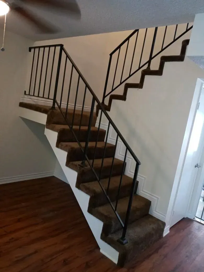 A set of carpeted stairs with a dark metal railing rising from a wood-floored room toward an upper landing.