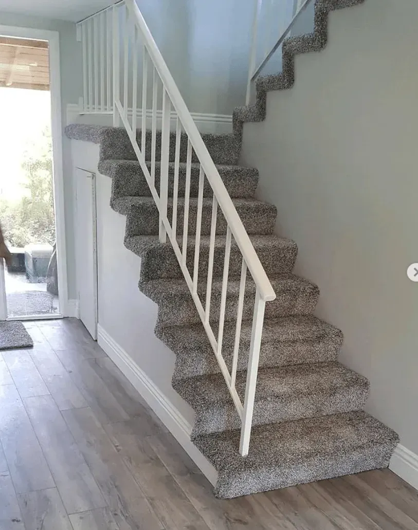 A carpeted staircase with white railings, leading upward in a bright interior with wood-look flooring.