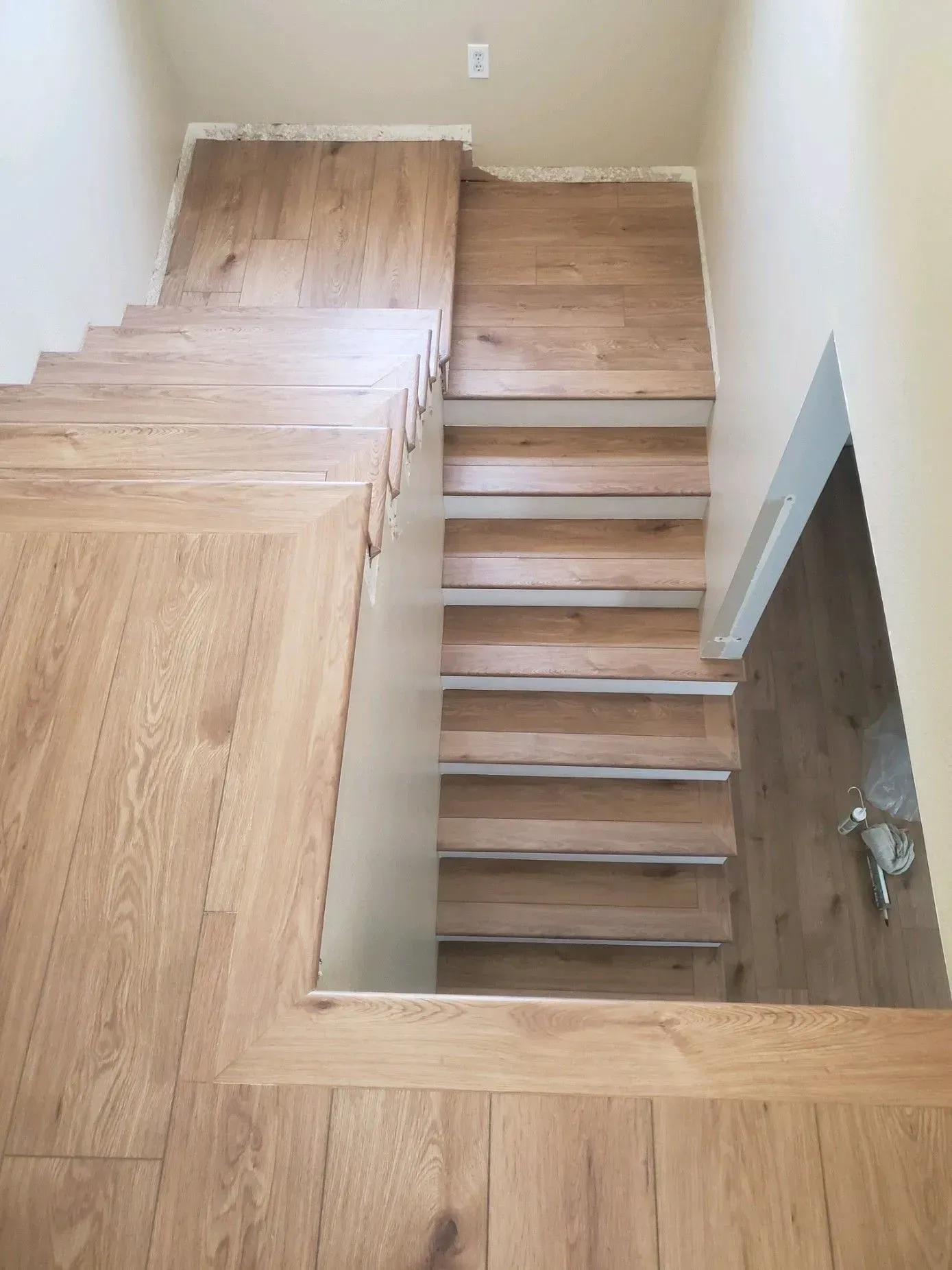 A high-angle view of a wooden staircase with a landing, leading down to a lower level with light-colored walls.