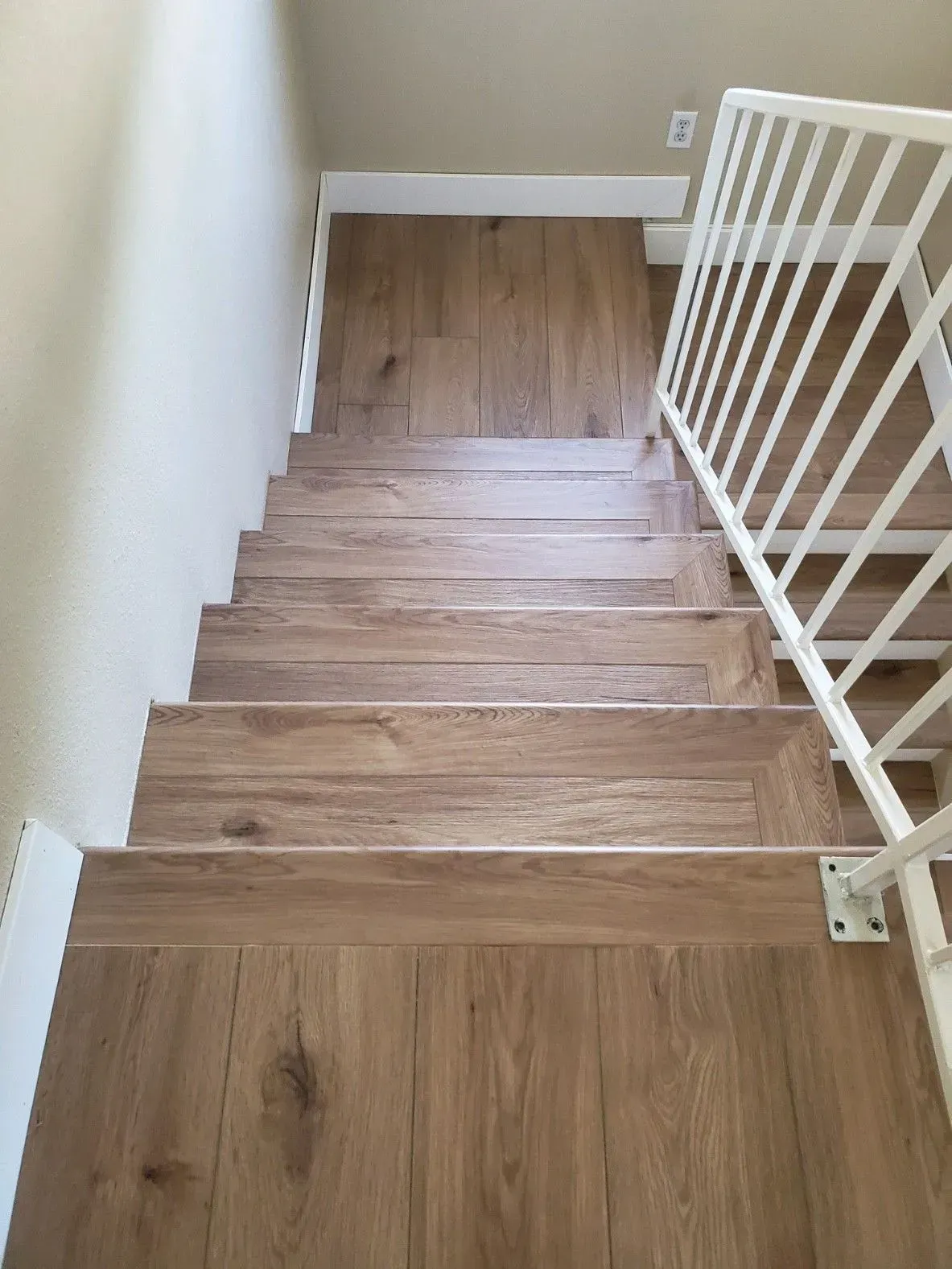 A top-down view of wooden stairs with light-brown planks, white trim, and a white metal railing.