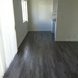 A bright, empty room with grey wood-look flooring, white walls, and a large sliding glass door with vertical blinds.