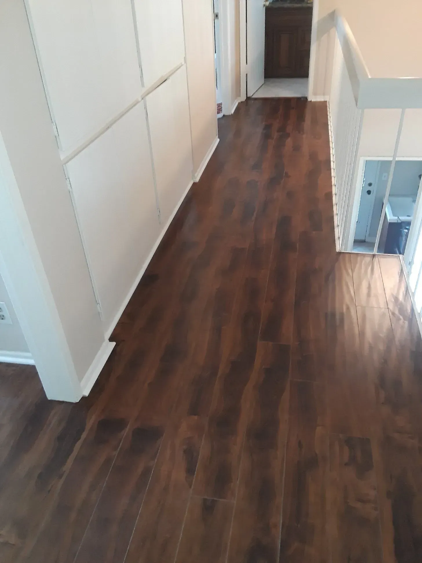 A hallway featuring dark brown wood-look flooring, light grey walls with paneling, and a staircase railing on the right.