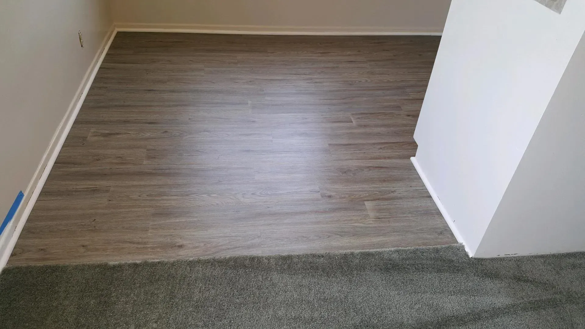 A transition between gray wood-look vinyl flooring and gray carpet in a room with white walls.