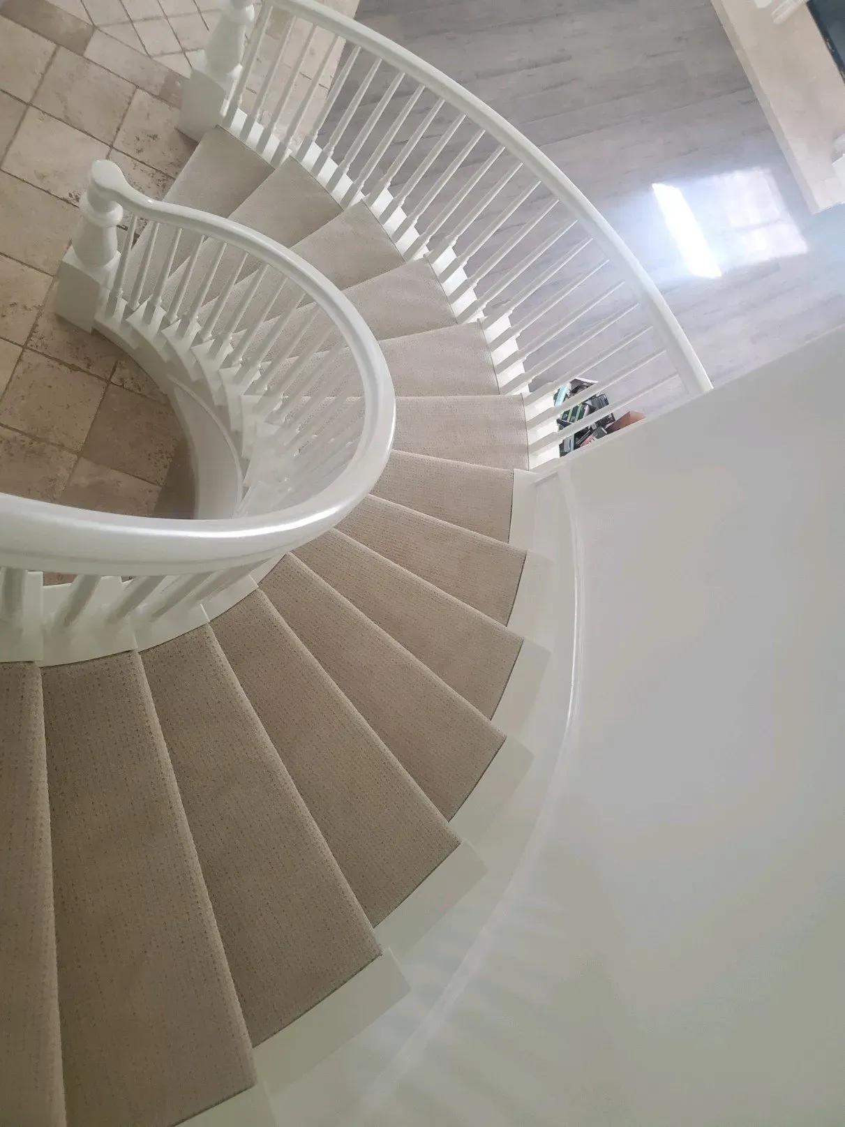A curved wooden staircase with white railings and beige carpet, viewed from above looking down into a tiled entryway.