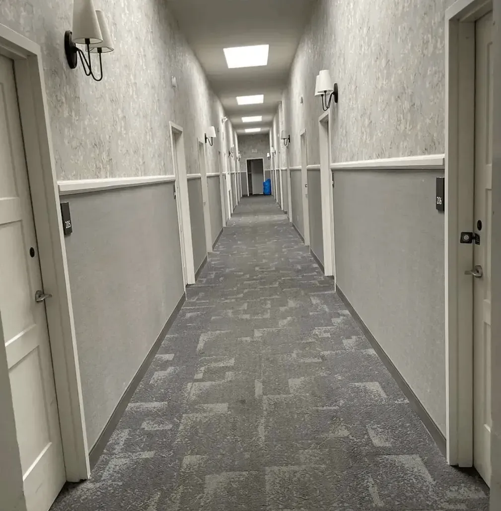 A long hotel hallway with grey carpet, light grey walls, white doors, and wall-mounted light fixtures.