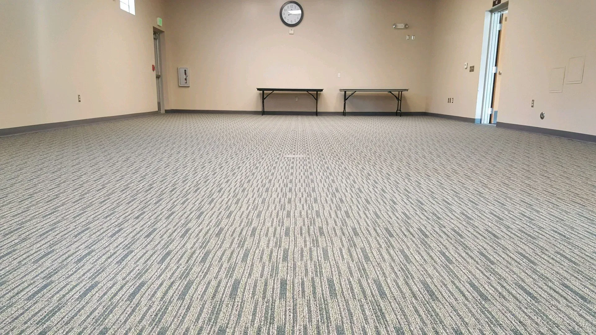 An empty community room with neutral-toned patterned carpet, two long tables against the back wall, and a round clock.