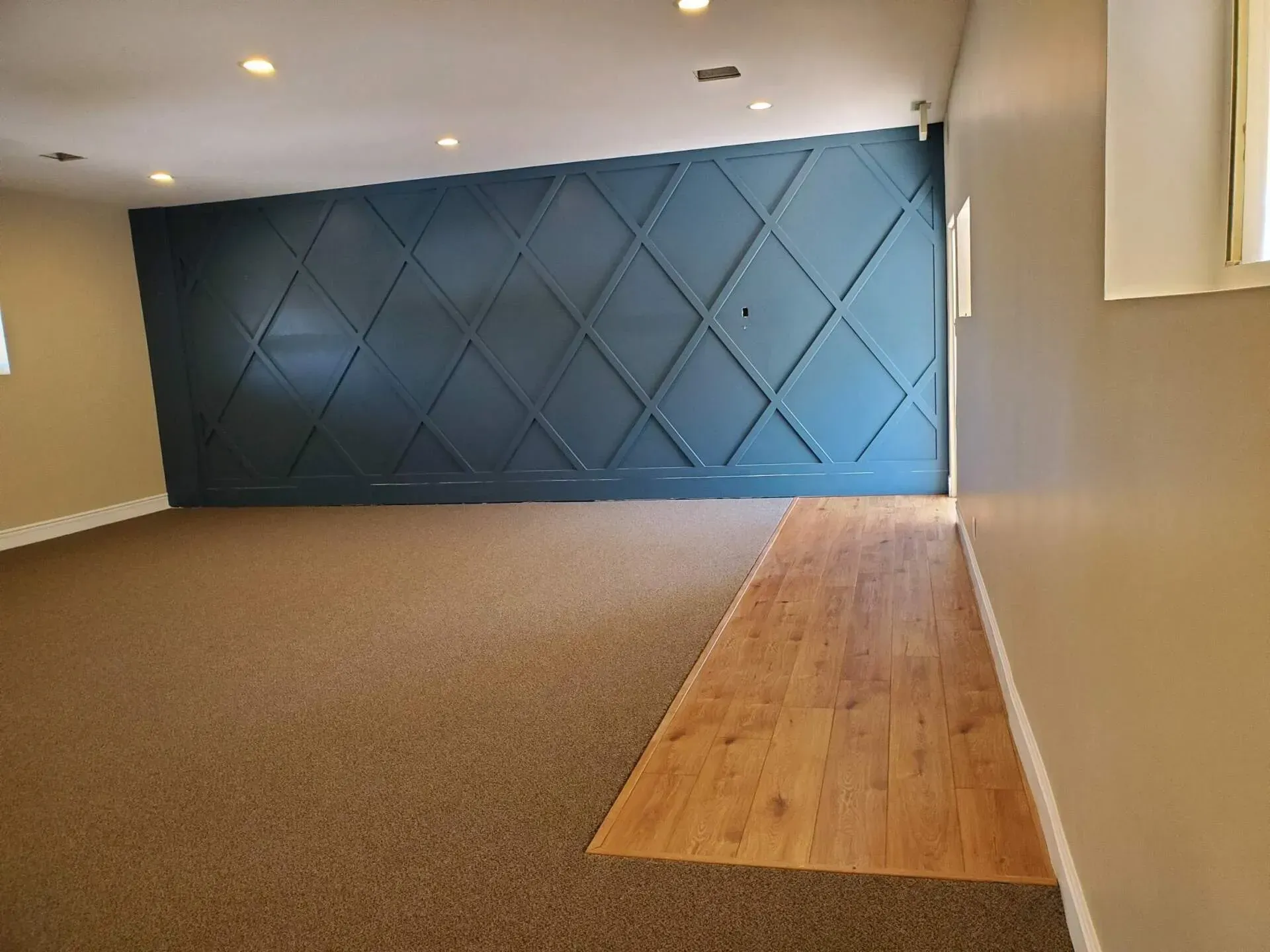 A room with brown carpet, light wood flooring, tan walls, and a large, dark blue accent wall featuring a diamond grid design.