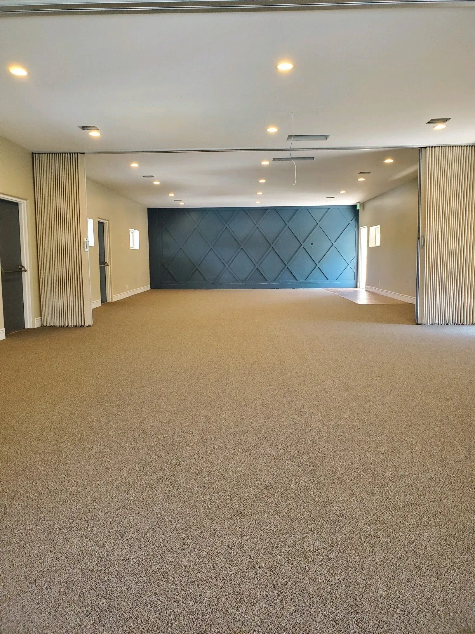 A large, empty event space with patterned beige carpet, cream walls, decorative curtains, and a dark blue accent wall.