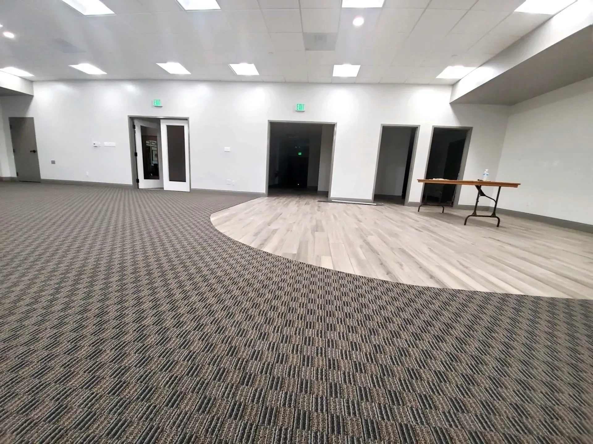 An expansive room with gray carpet, a light wood circular floor area, several doorways, and a single folding table.