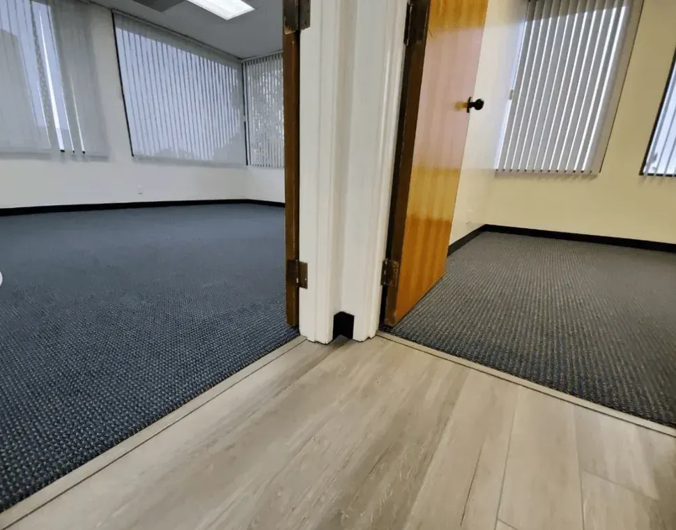 A wooden door frame separates two office rooms, both featuring gray carpet and vertical blinds, with light wood flooring.