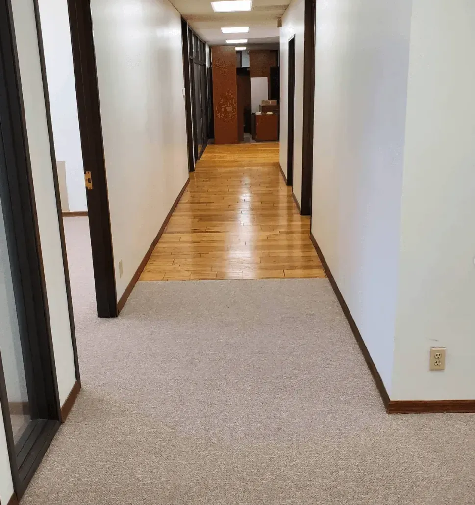 An indoor hallway transitioning from carpet to a wood floor, leading toward an open office space.