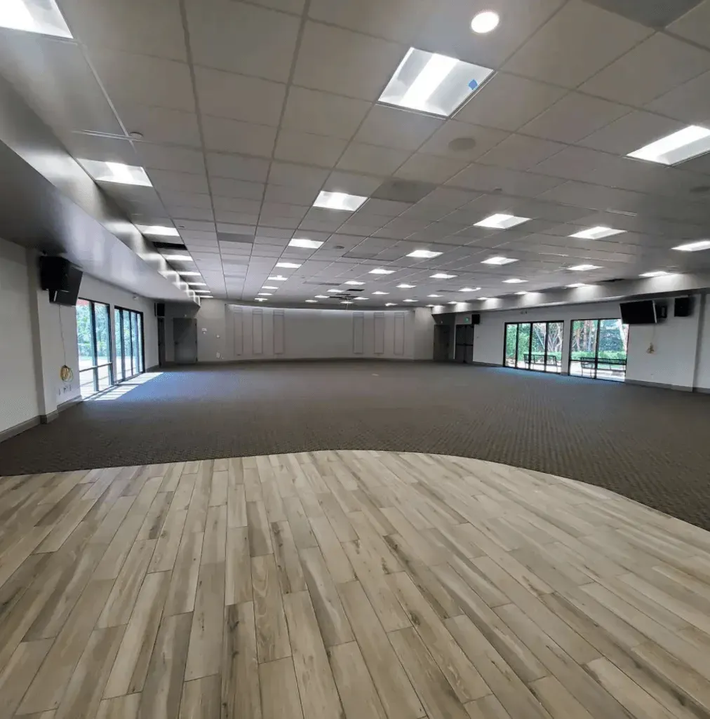 A large, empty event space with wood-look flooring in the foreground and gray carpet in the rear, featuring glass walls.