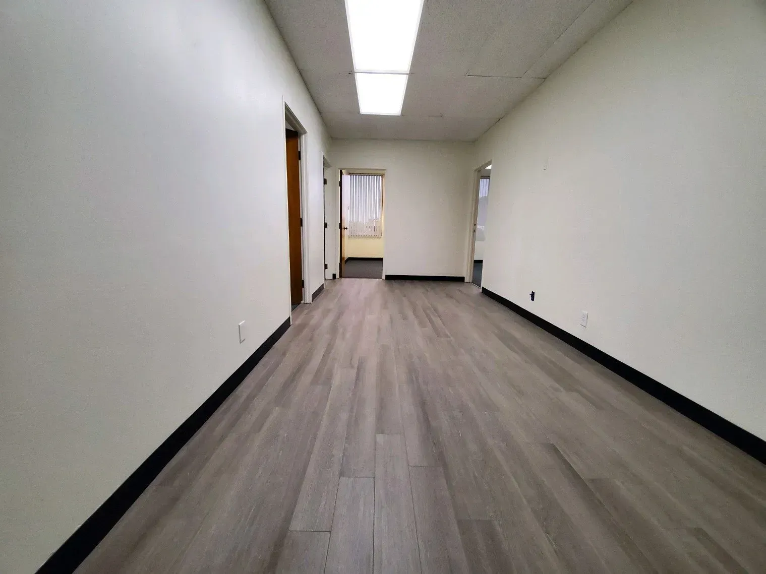 A long, empty commercial hallway with light walls, wood-look flooring, a drop ceiling, and doorways leading to other rooms.