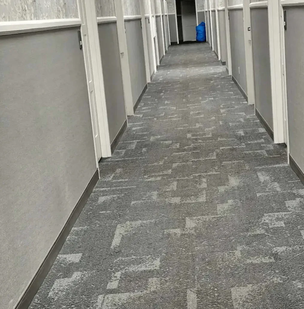 A long, empty indoor hallway with light-gray walls and patterned gray carpet, leading to a doorway with a blue item.