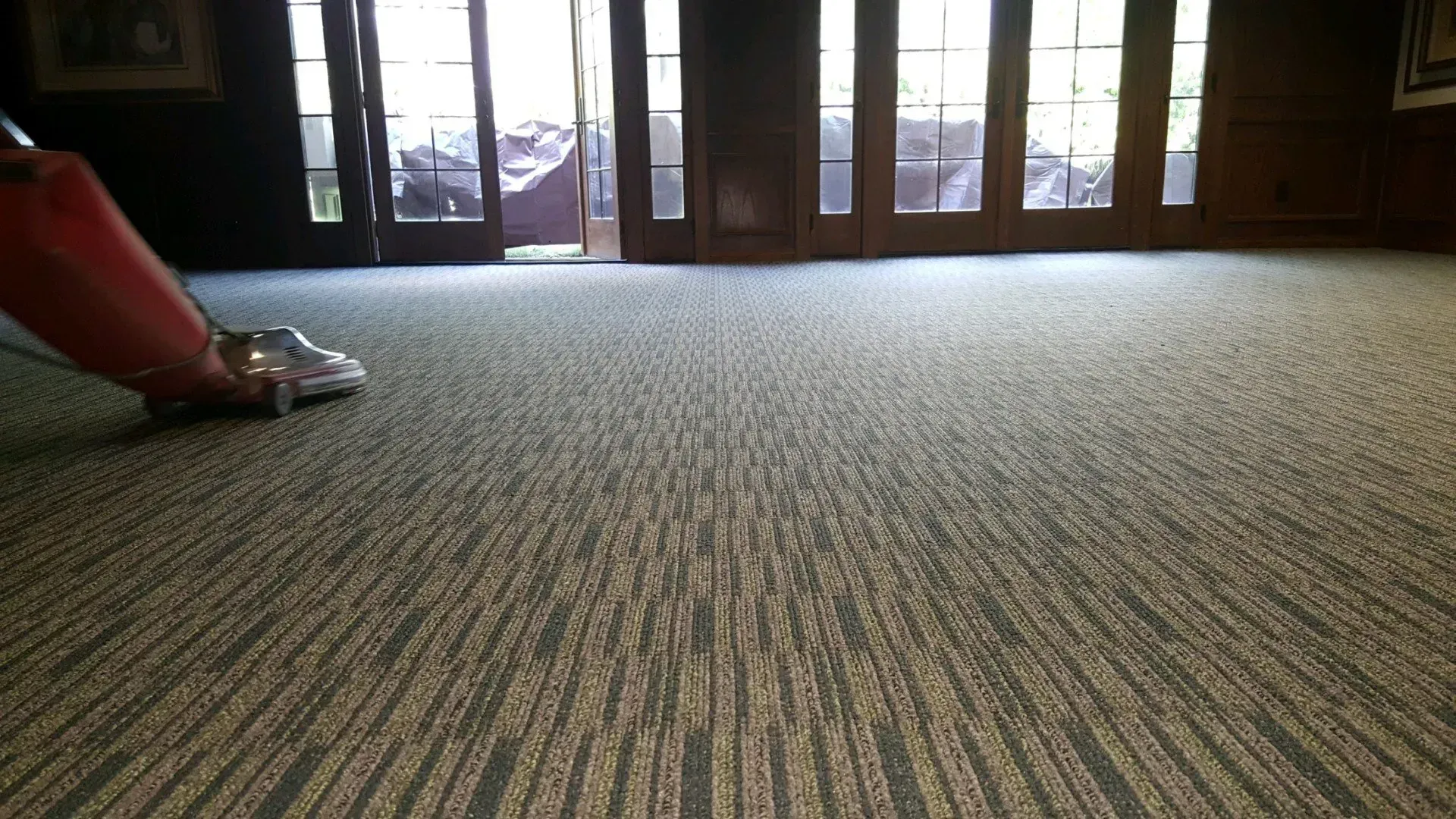 A person uses a red vacuum cleaner on a room's textured, striped carpet in front of glass doors.