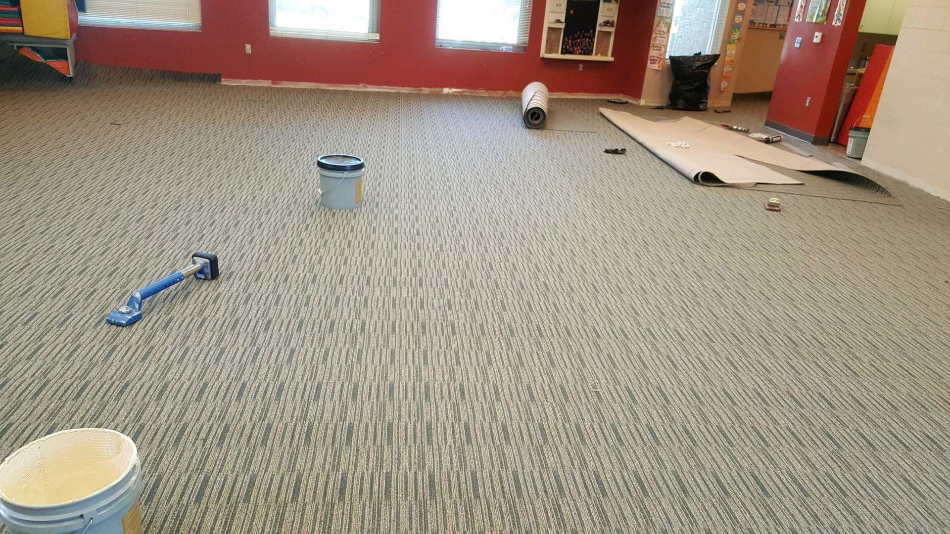 A room with a newly installed gray patterned carpet, featuring tools, a paint bucket, and rolls of flooring materials.