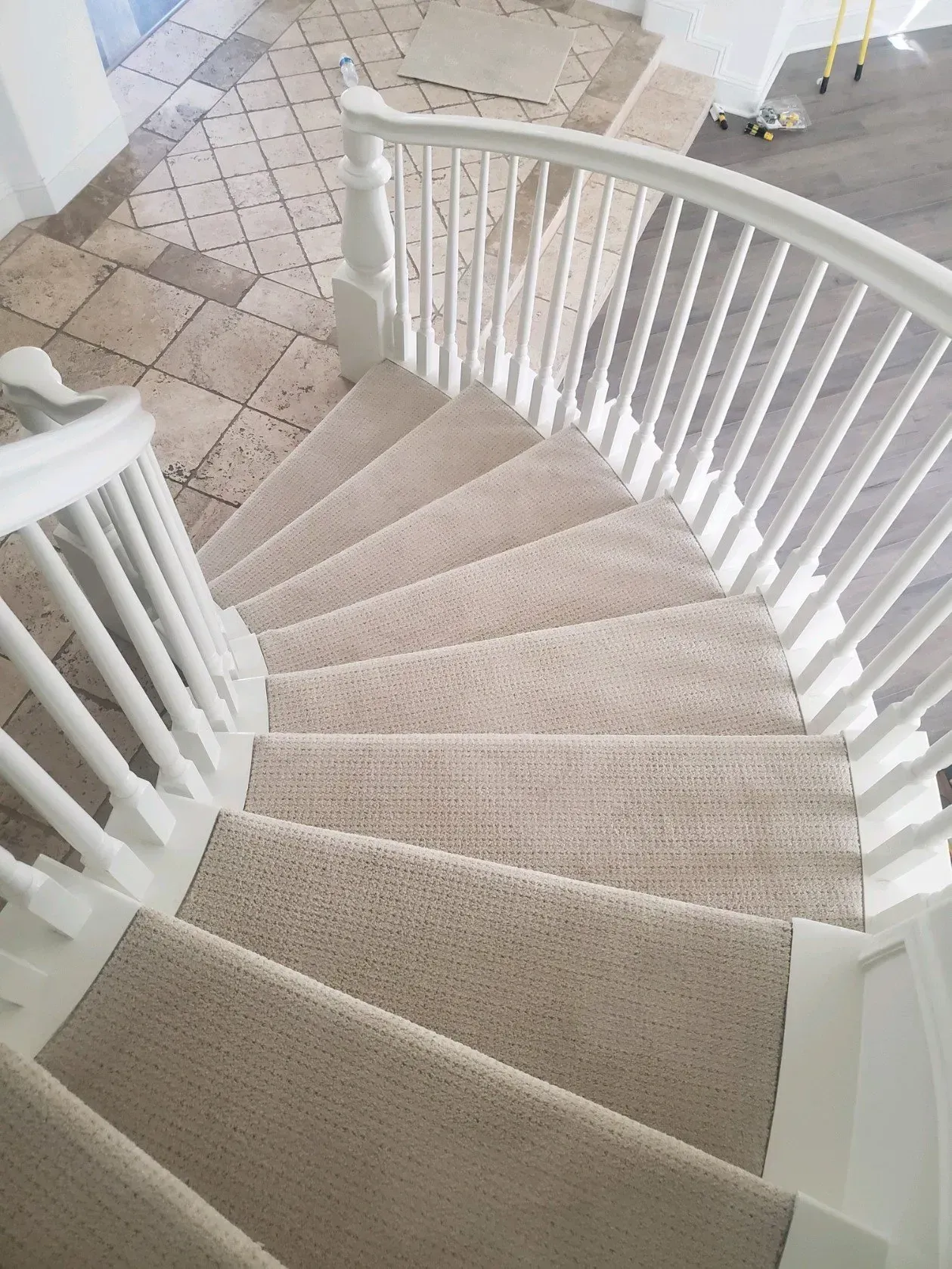 A curved staircase with beige carpet treads and white wooden railings, leading down to a tiled floor landing.