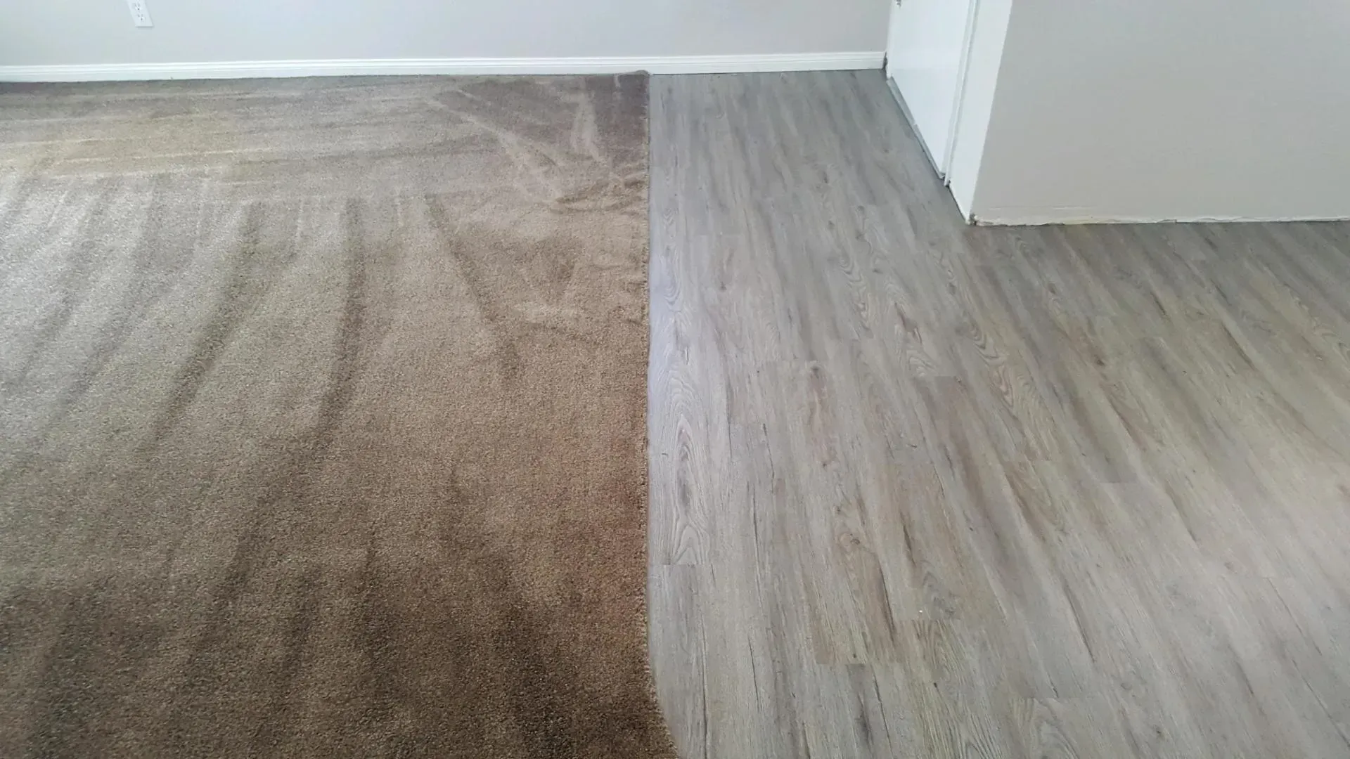 A room floor showing a transition between brown carpet and light gray wood-look vinyl planks.