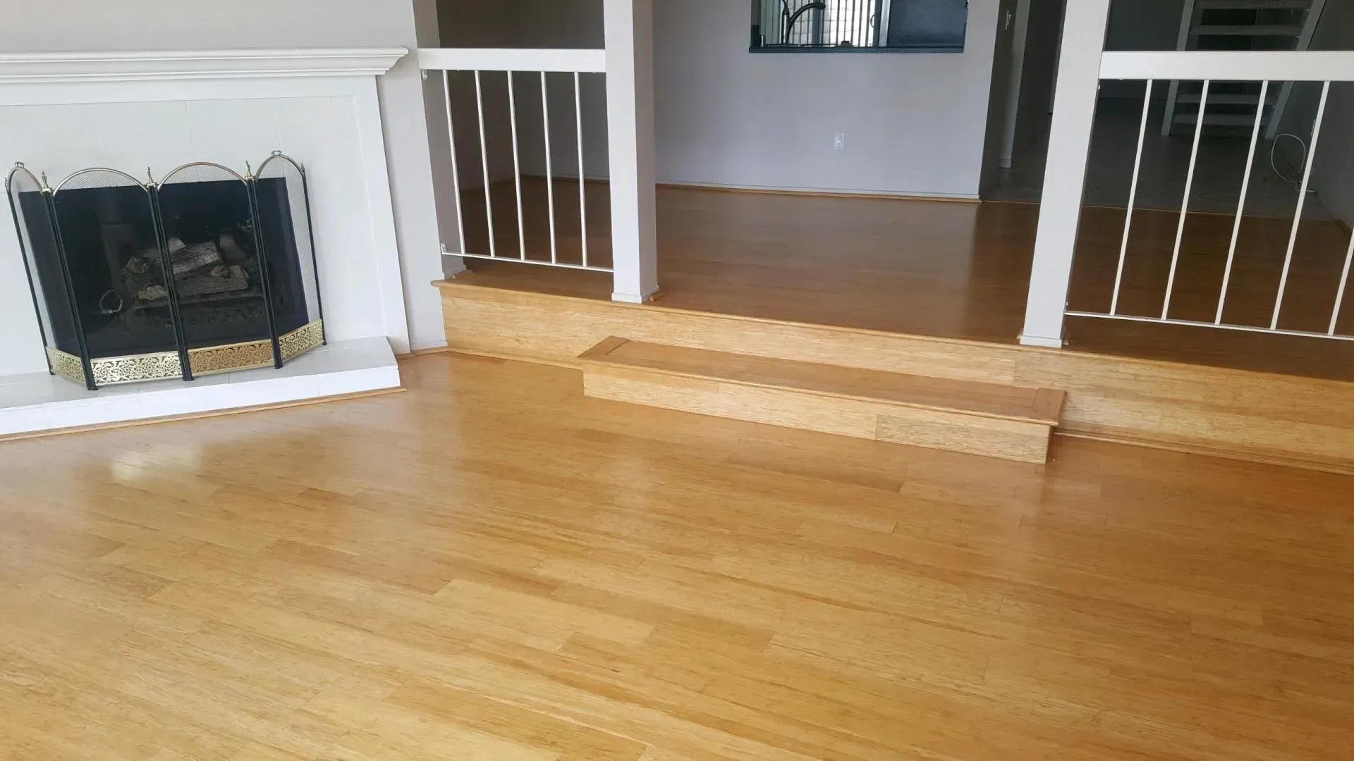 Light hardwood floors in a living room with a fireplace and a small step leading to an elevated area with railings.