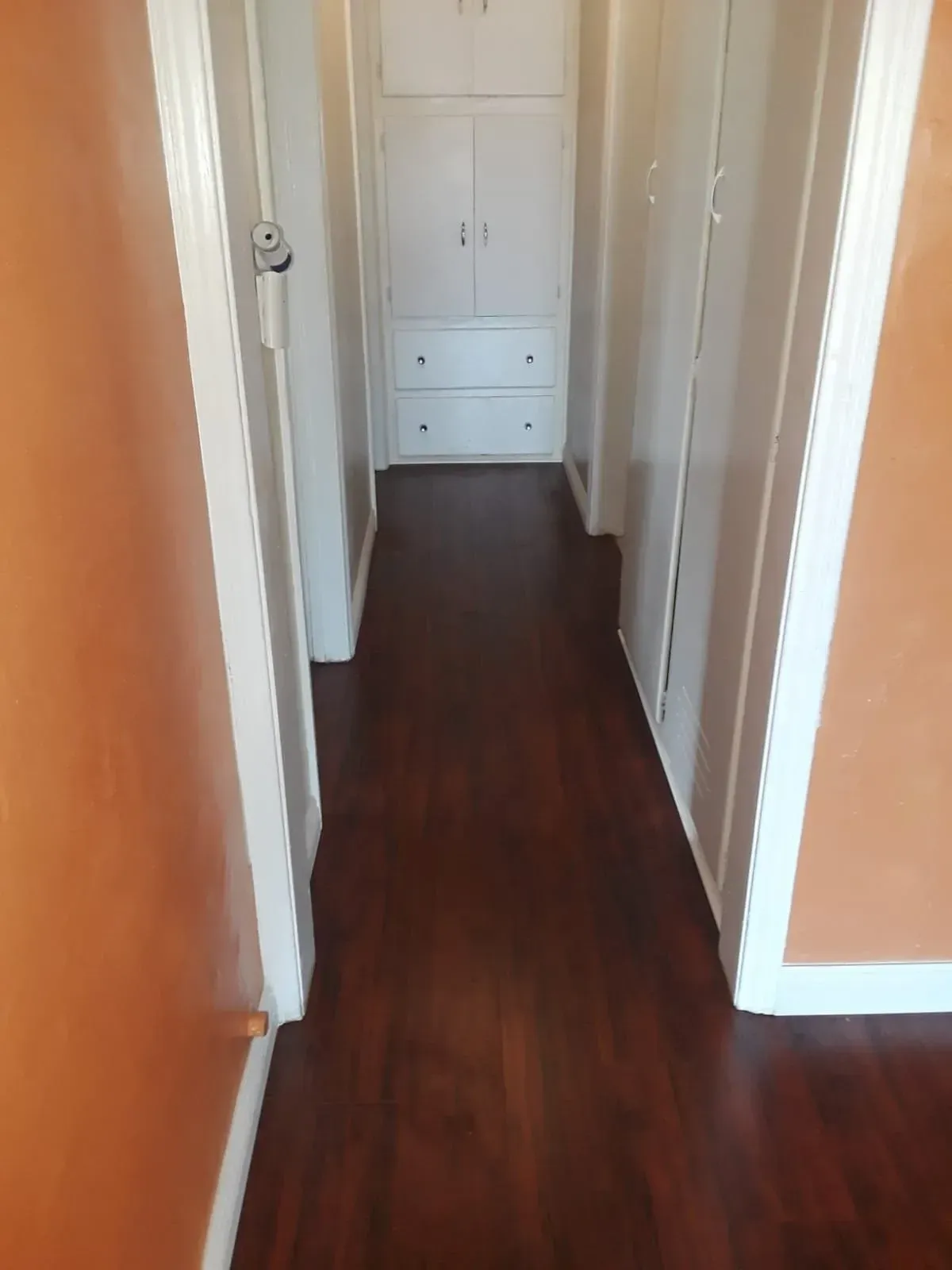 A hallway with dark wood flooring, orange walls, and white cabinets at the end.