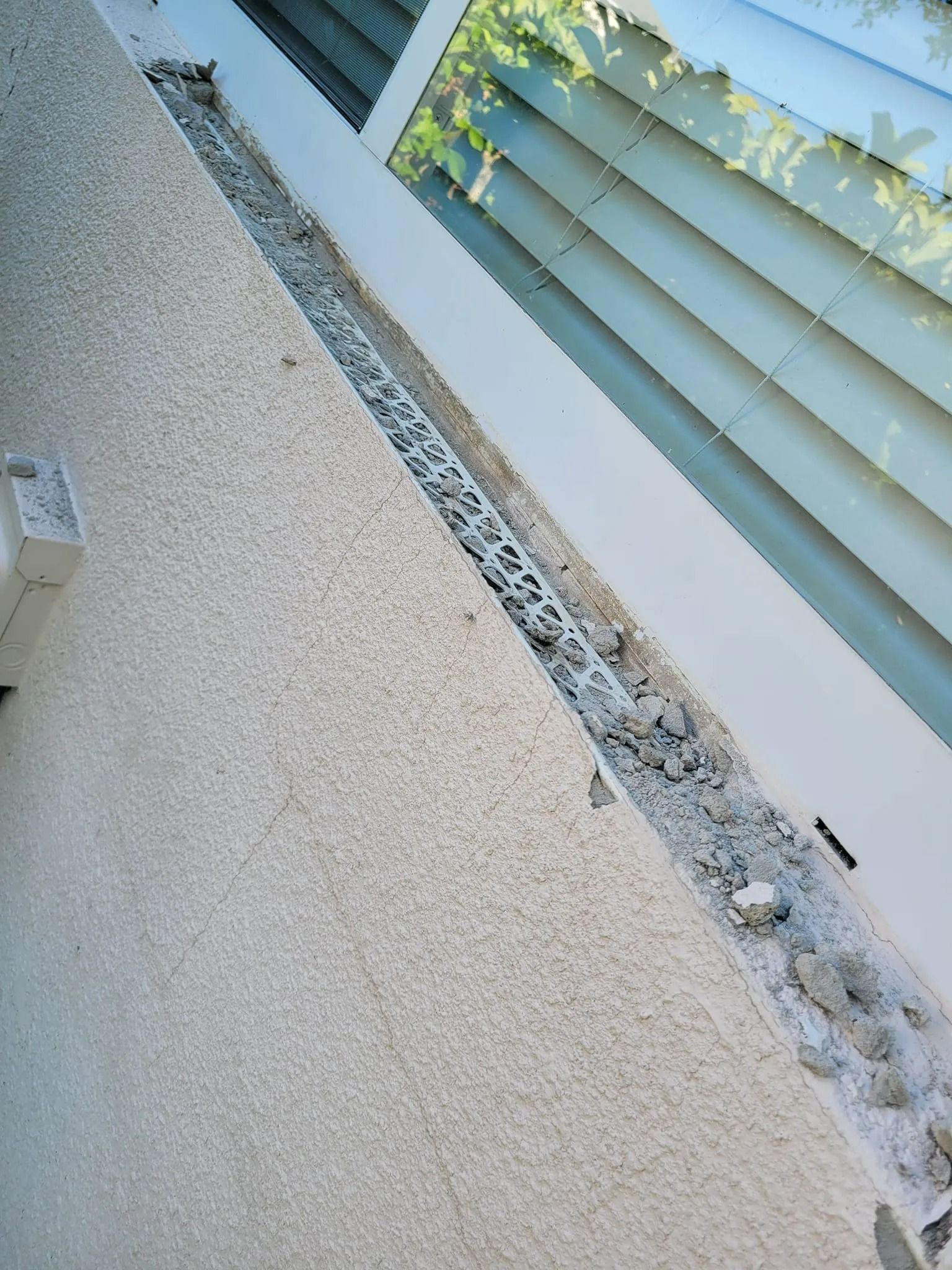 Exterior wall with a long cracked stucco seam under blue-gray window shutters and leaves above