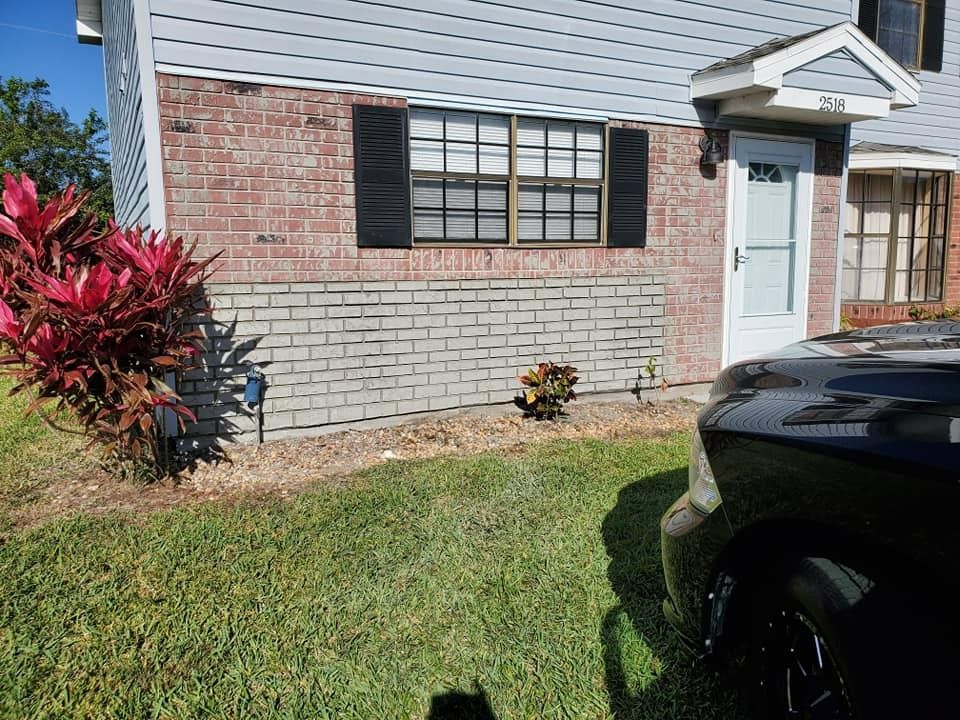 Front of a brick house with white siding, black shutters, small shrubs, and a car in the driveway