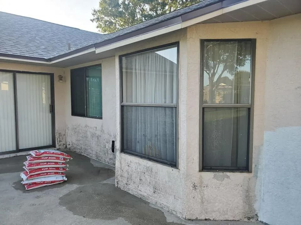Single-story house exterior with stucco walls, windows, and stacked red-and-white bags on the patio.