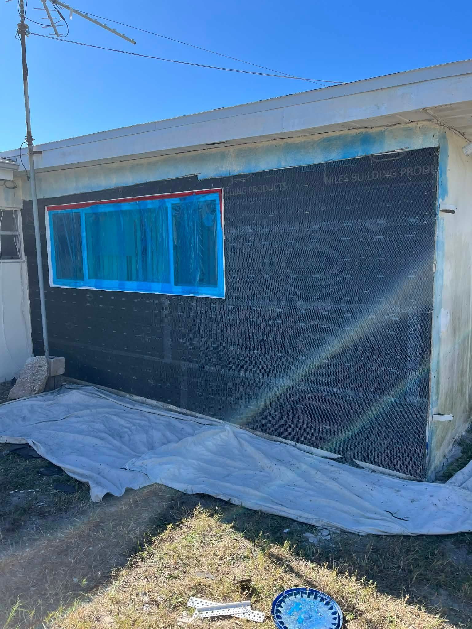 House exterior wrapped in black mesh with a blue-trimmed window and plastic sheeting on the ground