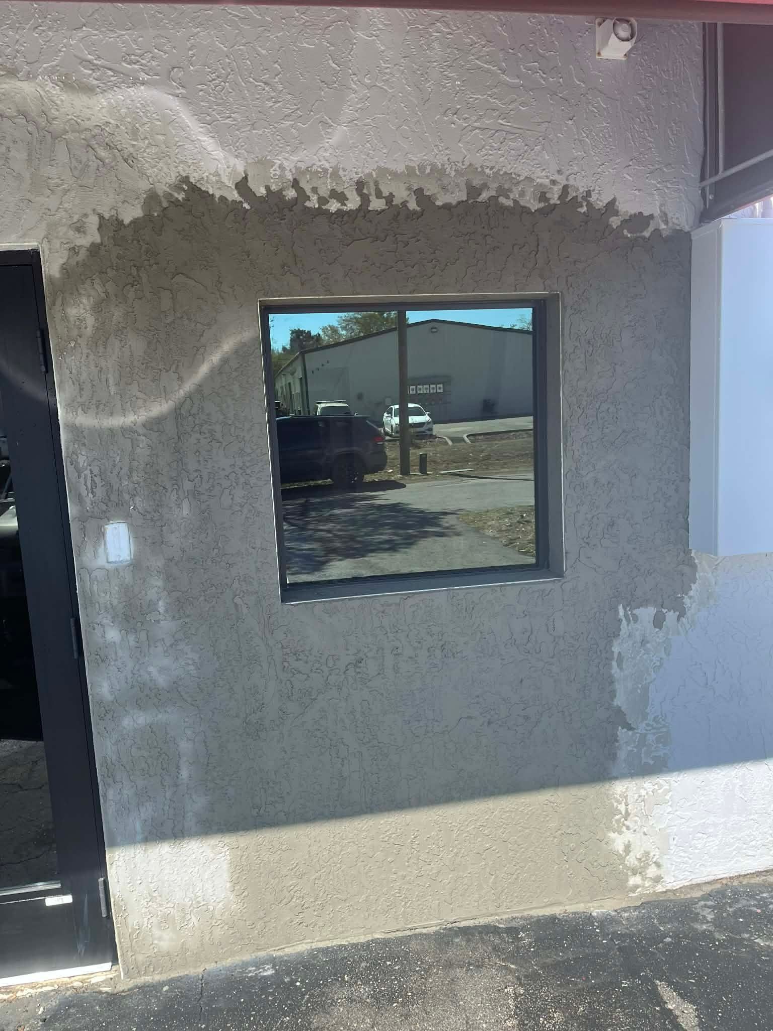 Stucco exterior wall with a dark-framed window reflecting a parking lot and building.