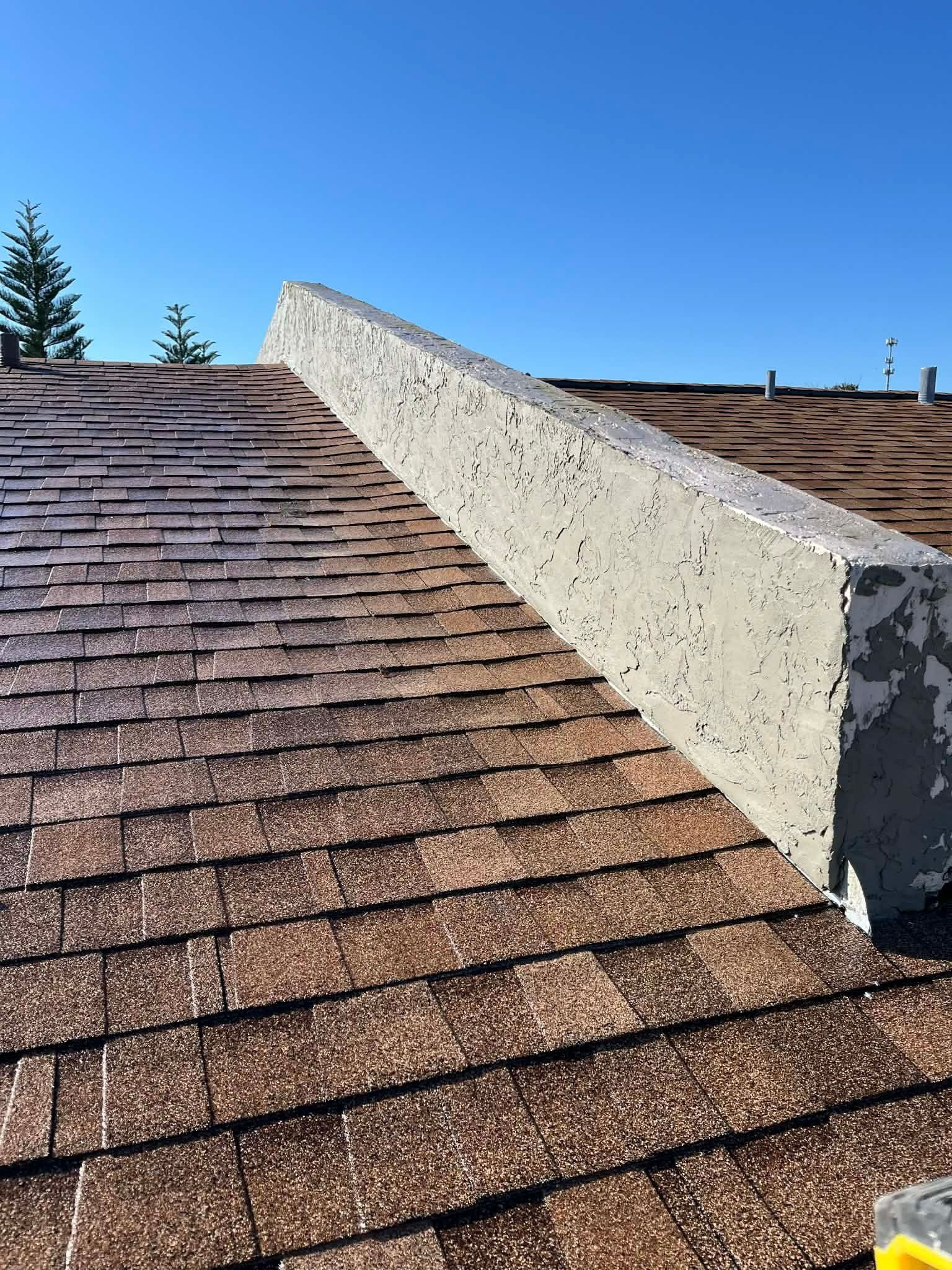 Asphalt shingle roof with a white chimney under a clear blue sky.