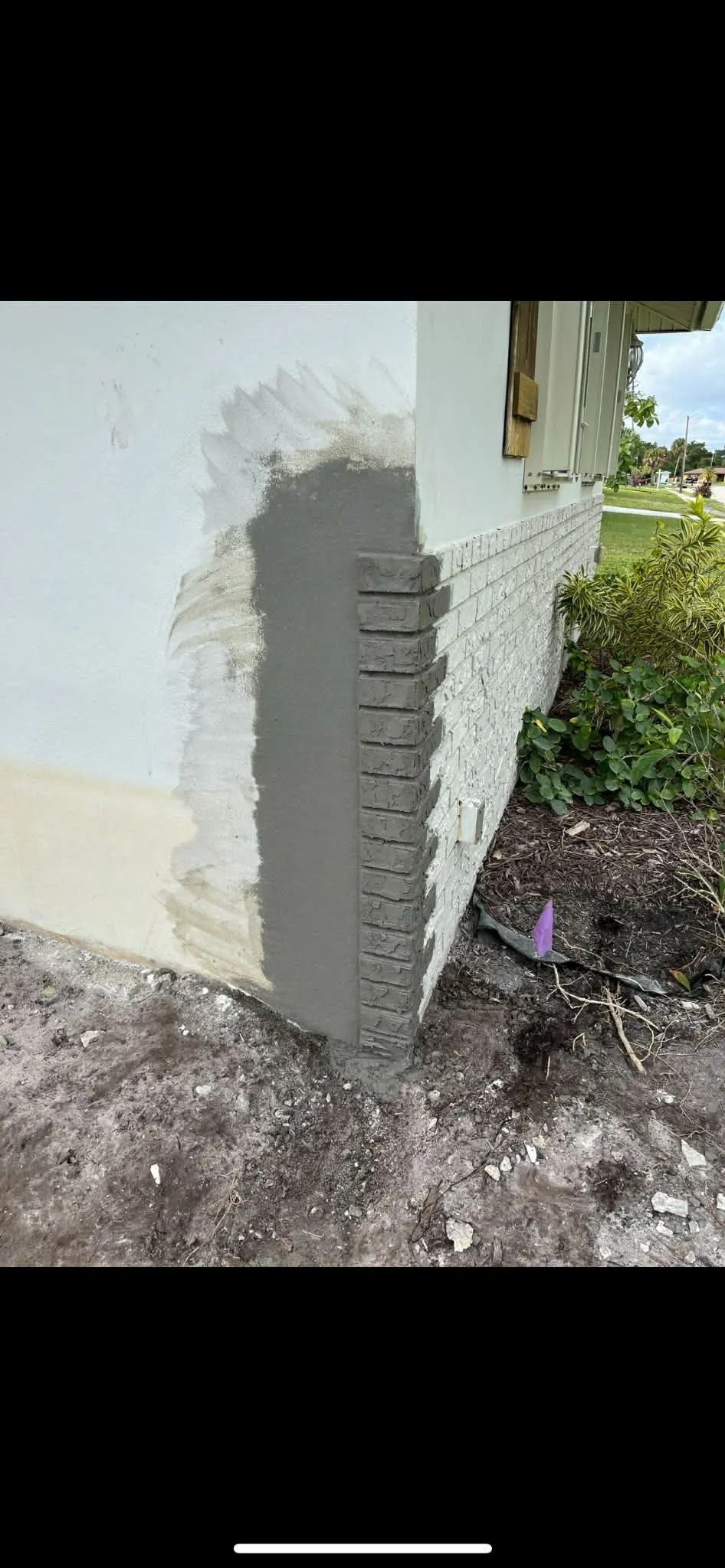 Close-up of a house corner with a white wall, exposed brick edge, and ground beside a garden path.